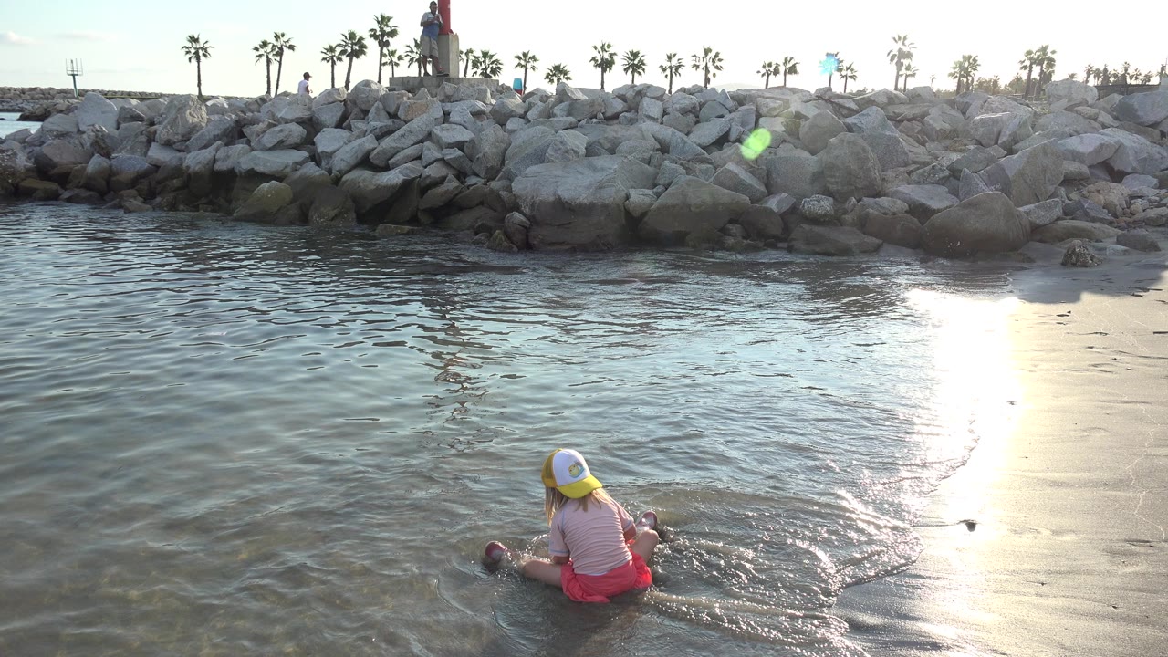 Young girl sits in shallow water near rocks and sun glare.