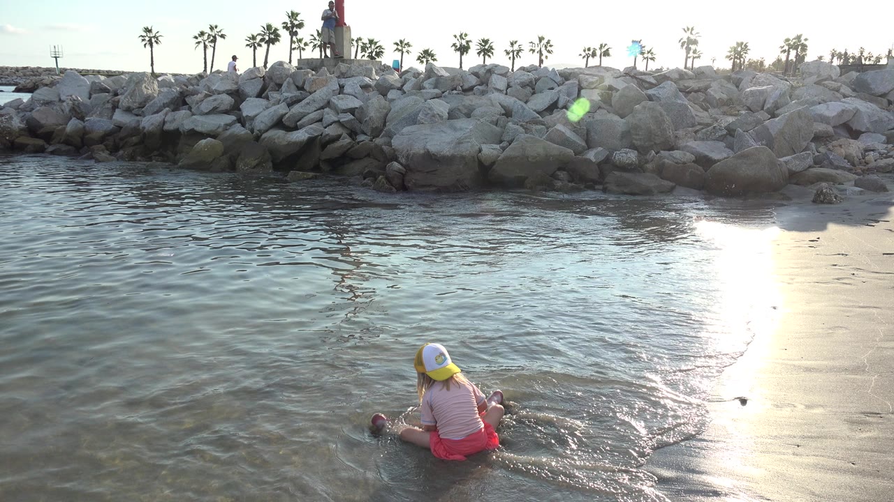 At 0:01, the girl remains seated in the shallow water in Grand Junction, Colorado, with small ripples around her legs and a strong vertical band of sun reflection on the right.