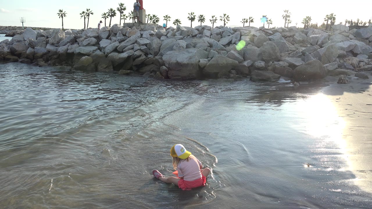 Young girl sits in shallow water near a sandy shore and rock breakwater.