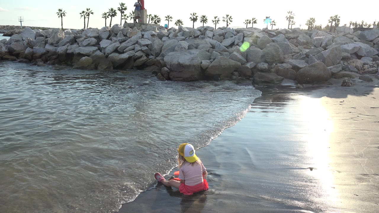 In Grand Junction, Colorado, a child in a yellow-and-white cap sits at the edge of shallow water while sun glare reflects on wet sand and rocks line the background.