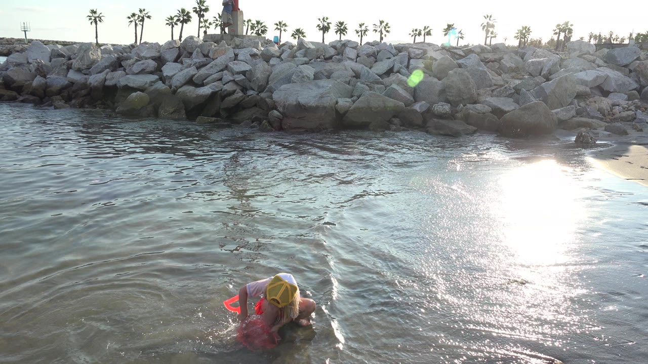At 0:00 in Grand Junction, Colorado, a child crouches low at the edge of shallow water with a red beach toy while sun glare reflects off the wet sand.