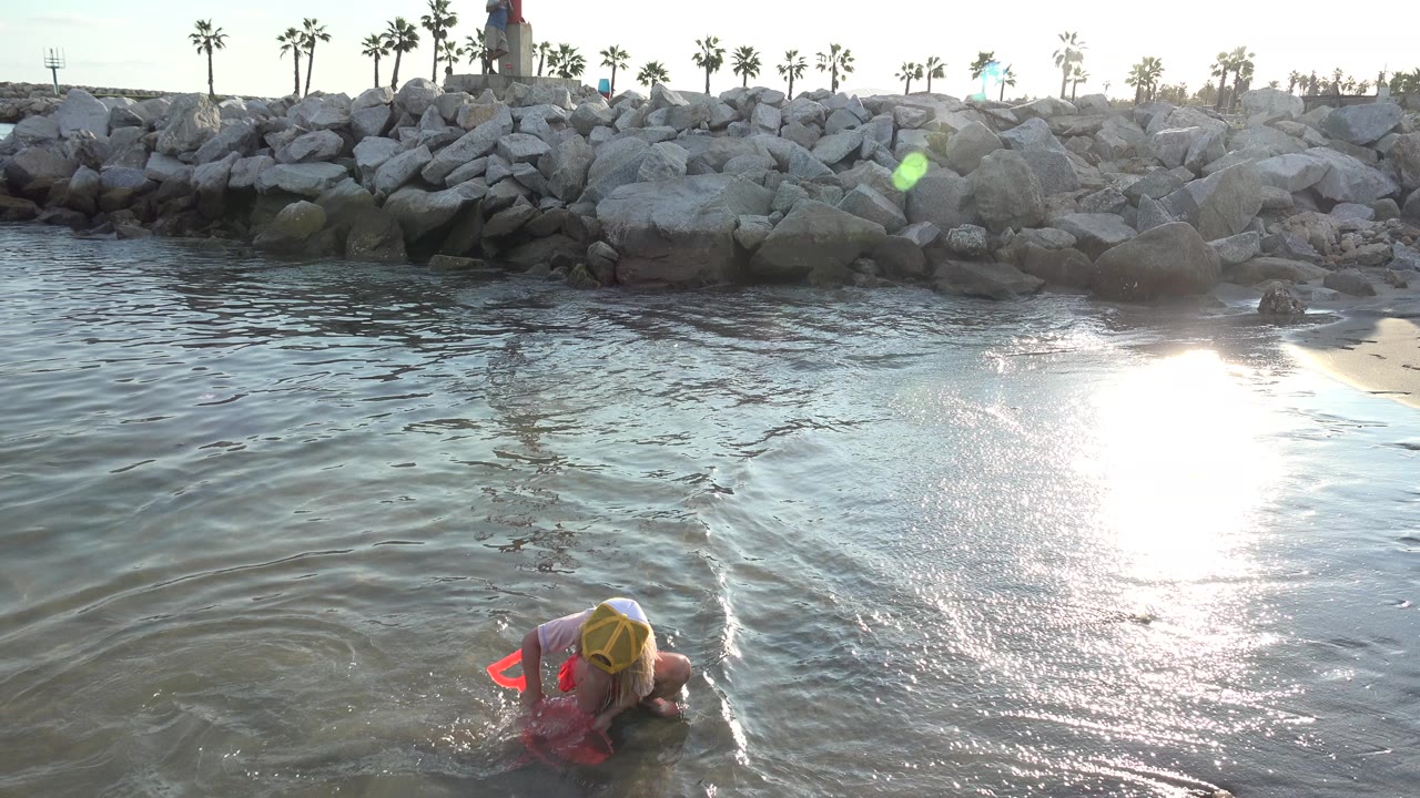 At 0:01 in Grand Junction, Colorado, the child remains near the lower edge as ripples move across shallow water and a bright reflection spreads over the sand.