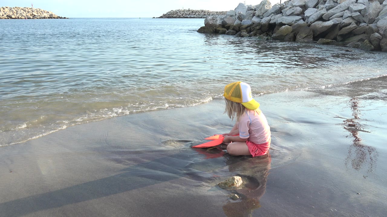 Little girl sits in wet sand beside shallow waves.
