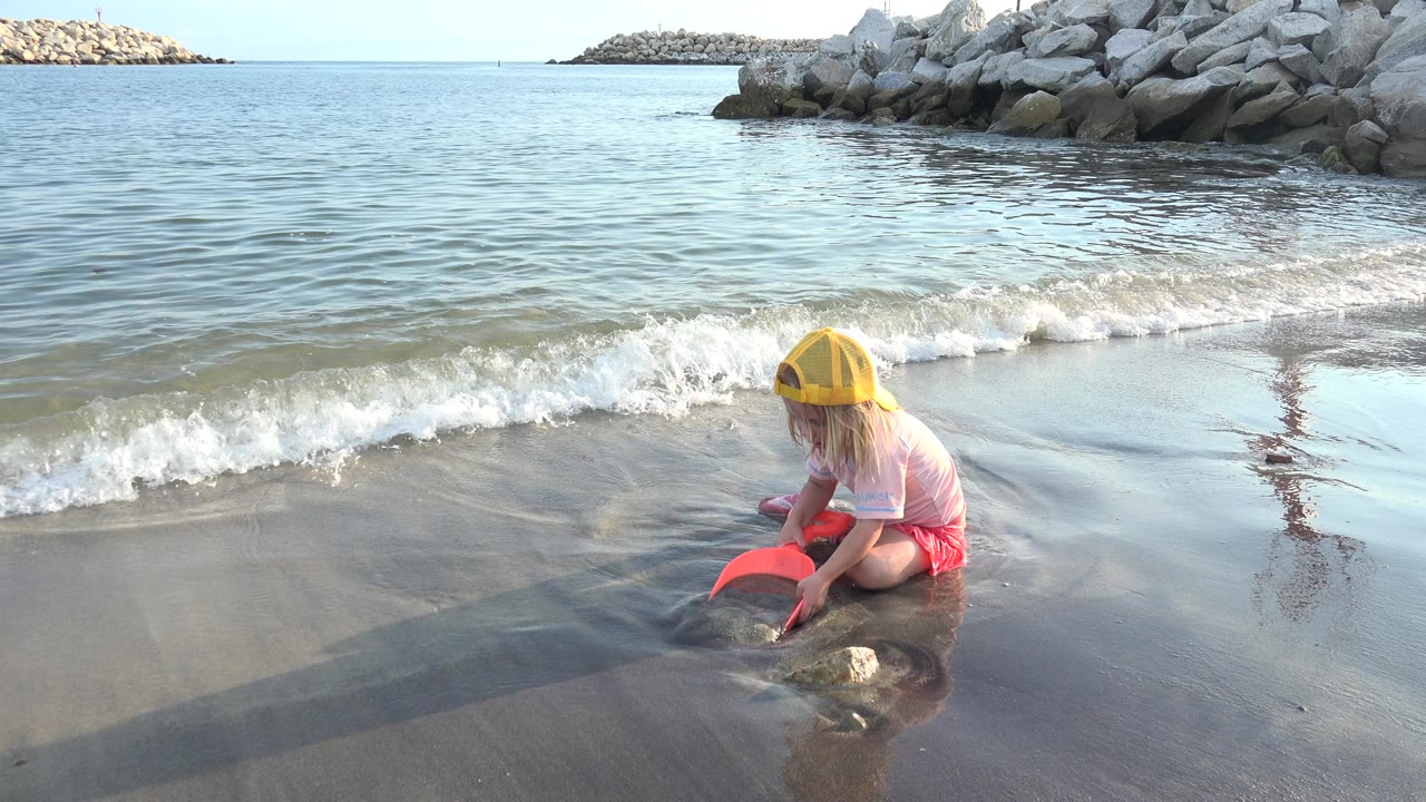 Young girl in a yellow cap playing at the wet shoreline with a red scoop.