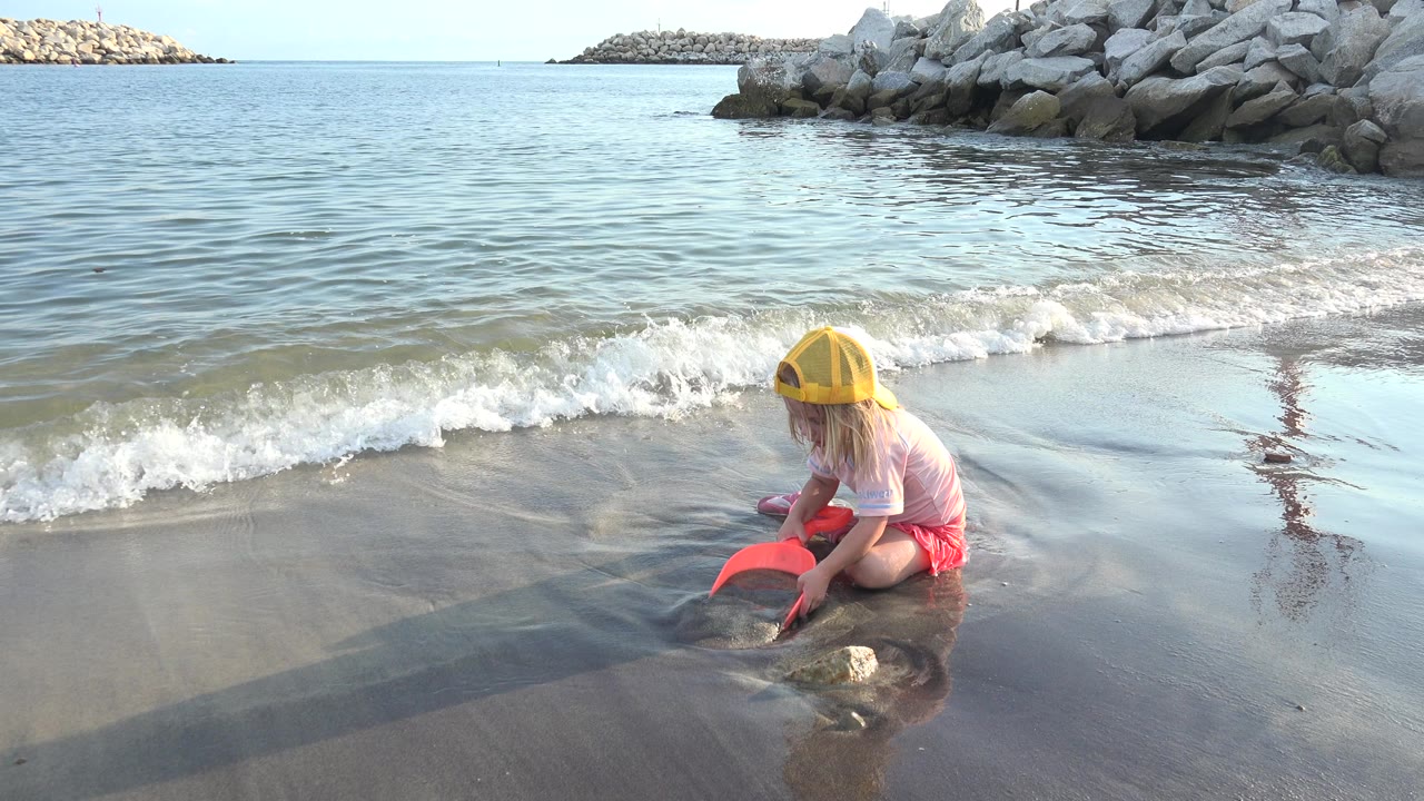 At 0:01 in Grand Junction, Colorado, the girl leans over the red scoop on reflective wet sand while a foamy wave line forms behind her.