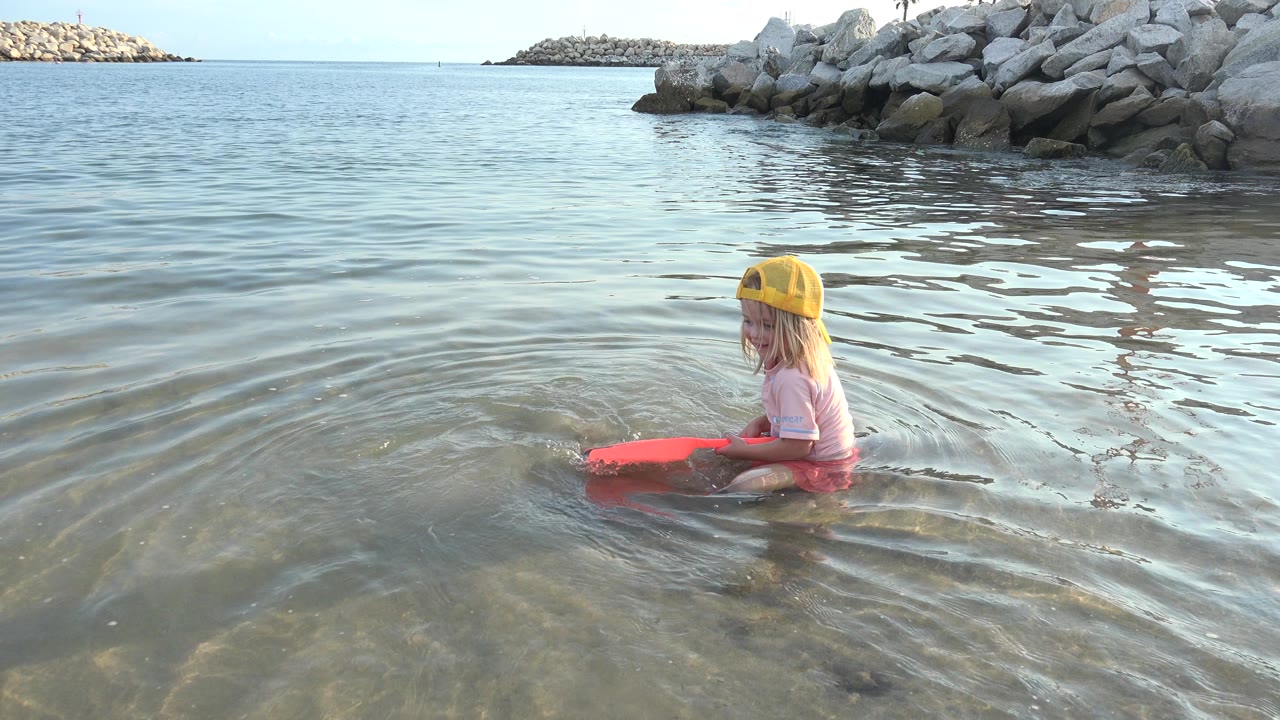 Young girl in a yellow cap sitting in shallow water with an orange scoop.