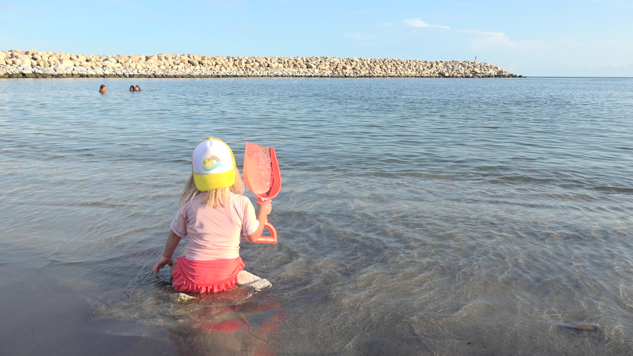 Blond child sitting in shallow water with a red scoop near a calm shoreline.
