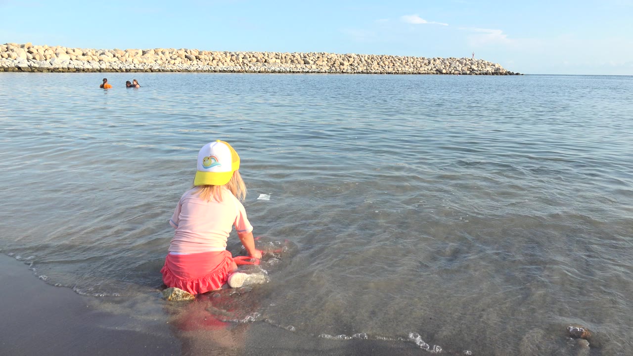 In Grand Junction, Colorado, a young girl in a yellow-and-white cap sits near the waterline as shallow waves reach her legs and distant swimmers appear near the breakwater.