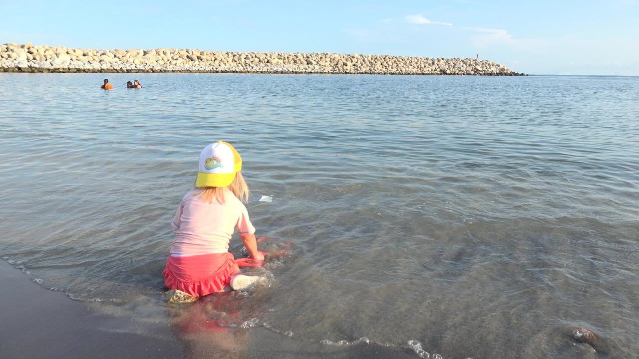 In Grand Junction, Colorado, the girl remains low in the shallows facing away from camera, with ripples spreading around her and two swimmers small in the background.