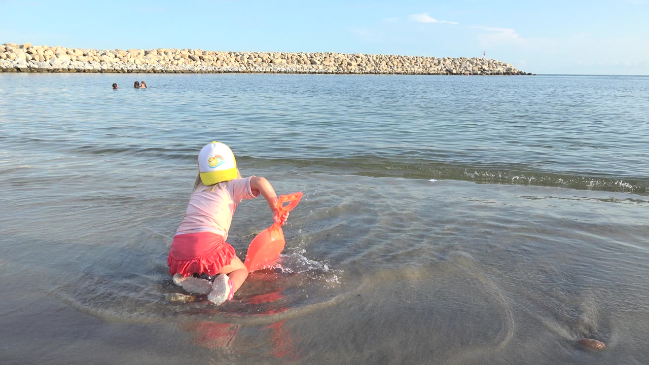 Young girl in a yellow and white cap kneels in shallow shoreline water in Grand Junction, Colorado, holding an orange sand toy as small ripples move around her.