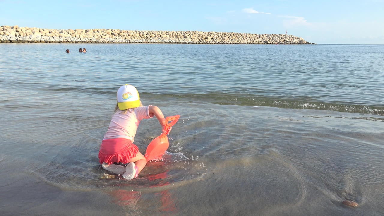Child in a cap leans over an orange sand toy at the Grand Junction shoreline while a small splash spreads across the wet sand in front of open water.