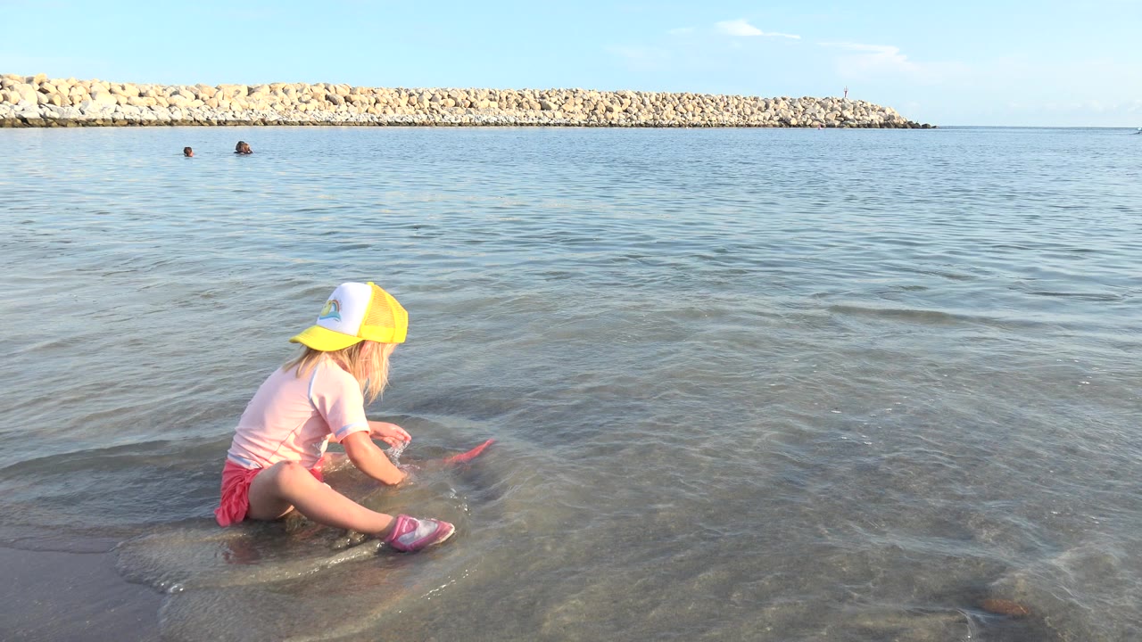 At 0:00 in Grand Junction, Colorado, a blonde child in a yellow-and-white cap sits near the left edge of shallow water holding a red scoop toy, with a rock breakwater and two distant swimmers behind.
