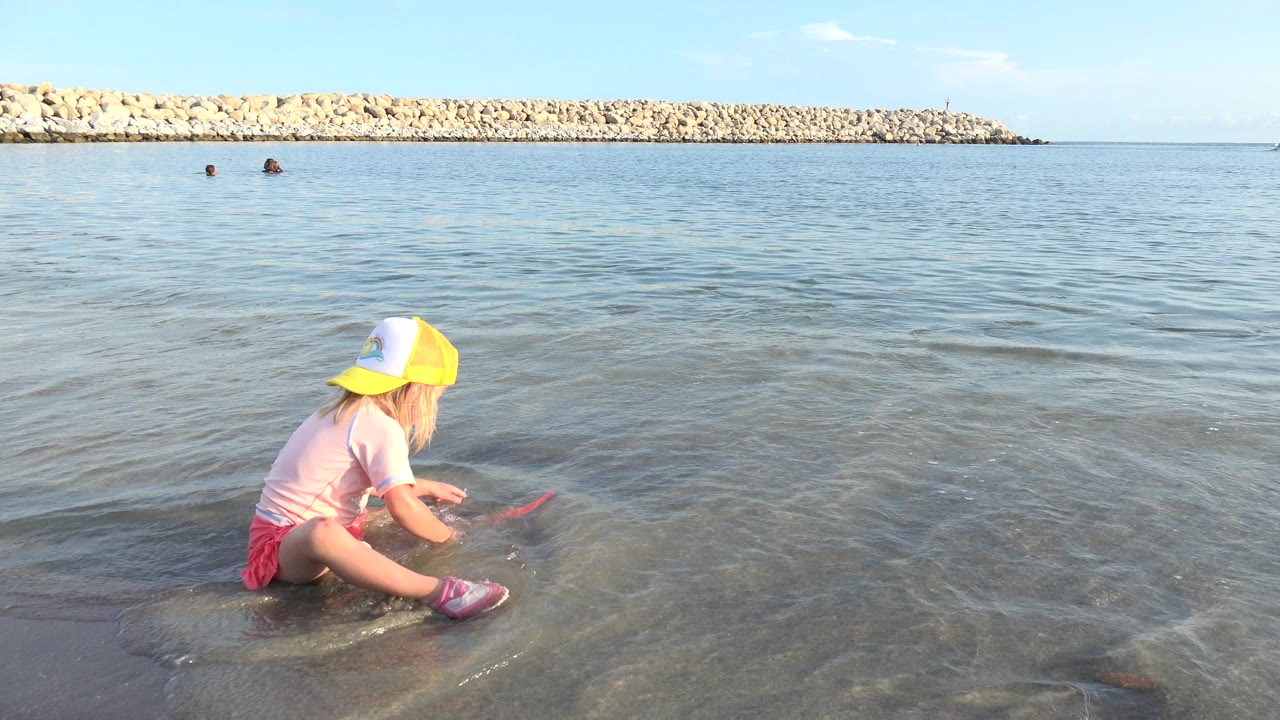 At 0:01 in Grand Junction, Colorado, the child leans forward over the red scoop toy as thin waves cross the wet sand, with the long rock breakwater steady on the horizon.