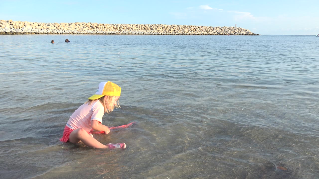 Child sits lower in the shallow water while holding the red toy.