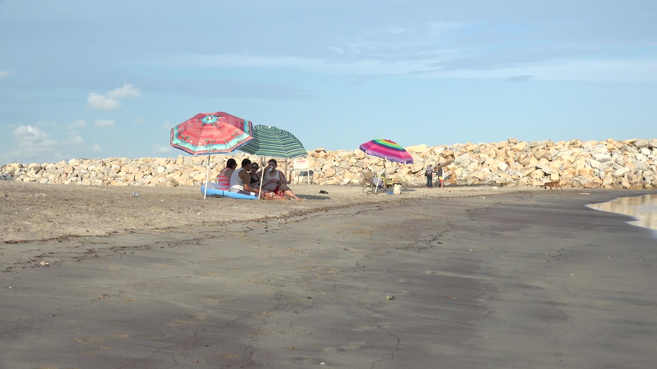 Beach umbrellas and seated people on wet sand beside rocks and blue sky.