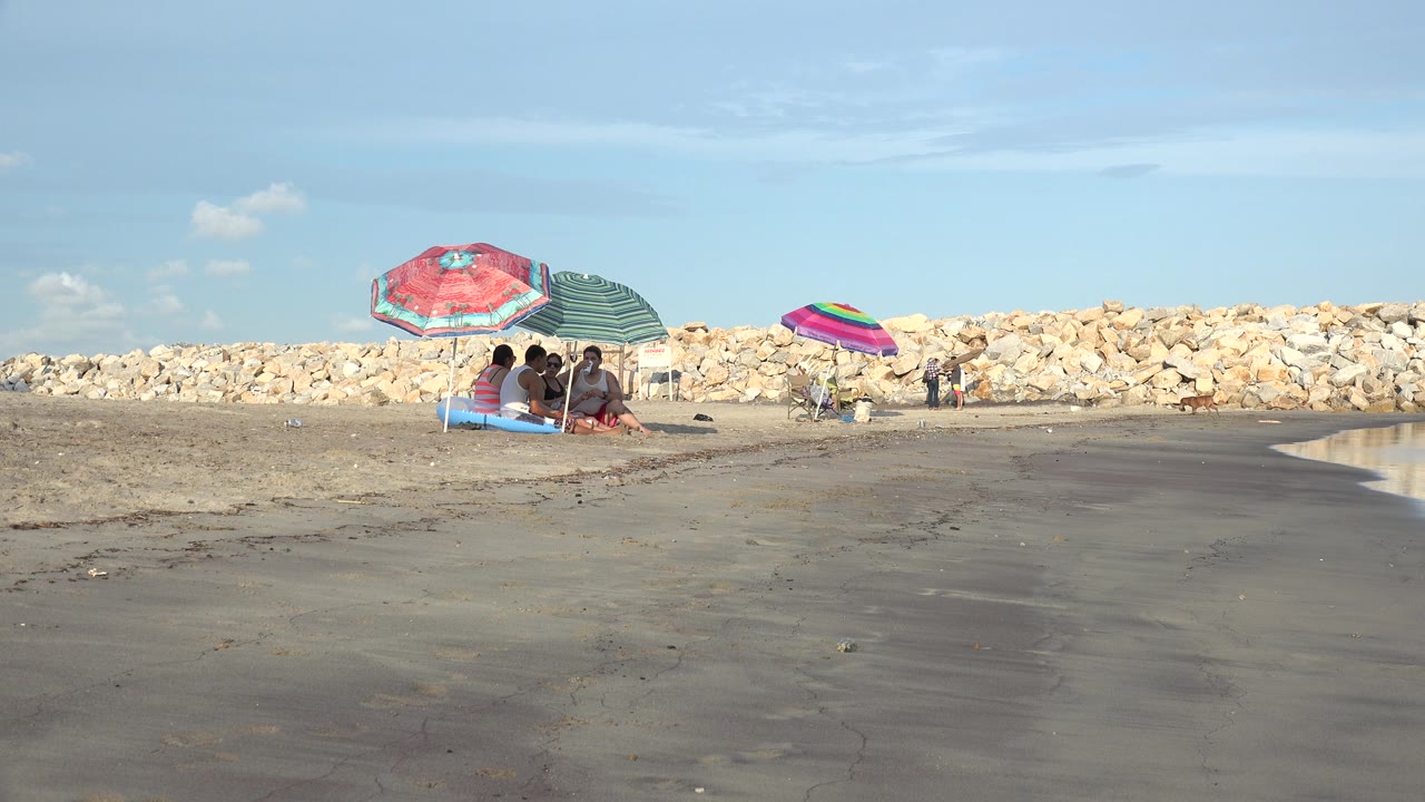 Grand Junction, Colorado beach frame with the seated group still near the left under umbrellas, open wet sand in front, and a narrow waterline entering at the right.