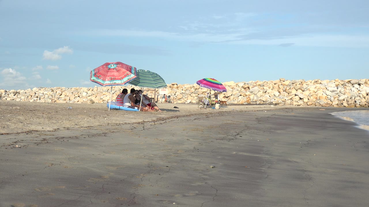Opening frame of the Grand Junction, Colorado beach scene, with seated people under red and green umbrellas at left and a striped umbrella farther back near the rocks.