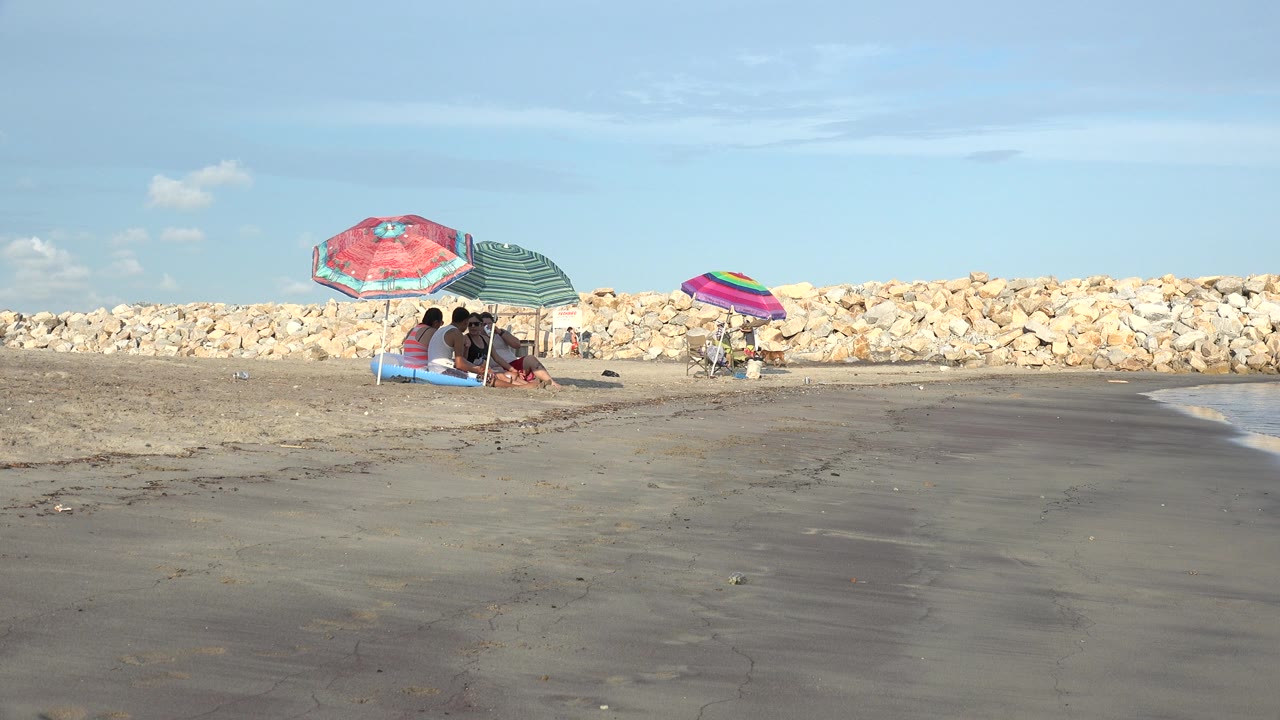 Early frame from the Grand Junction, Colorado clip showing the seated group under two umbrellas at left, the multicolor umbrella nearer center, and a darker wet-sand band crossing the foreground.