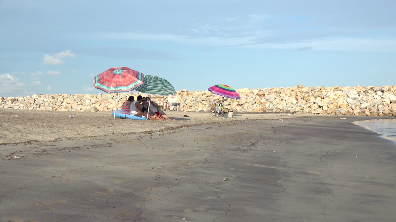 Small group under umbrellas on a mostly empty beach with rocks behind them.