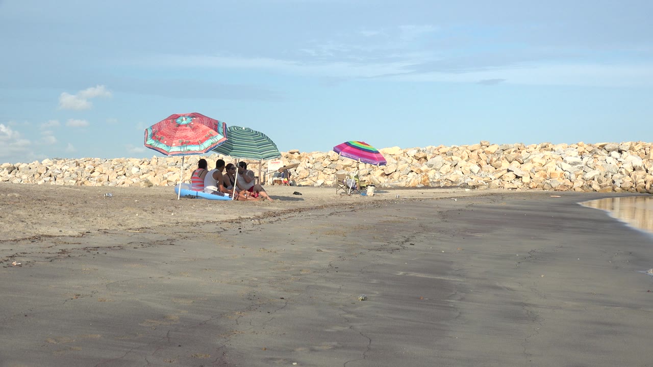 At 0:00 in Grand Junction, Colorado, people sit under red and green umbrellas near the shoreline, with wet sand in front and a pale rock barrier behind them.