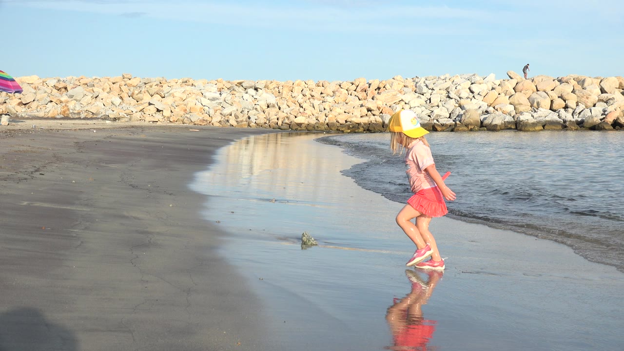 Young girl in a yellow hat walking on wet sand with a red beach shovel.