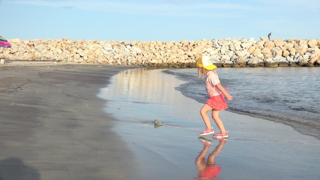 The girl continues along the Grand Junction, Colorado shoreline, holding the red shovel as shallow water curls beside the reflective sand.