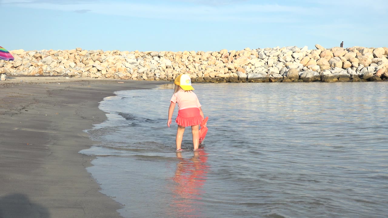 Child standing in shallow water at a beach with a rock barrier behind.