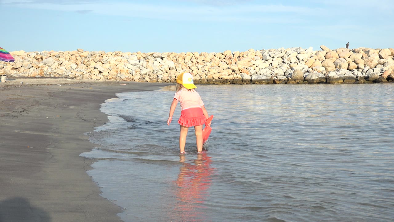 The girl remains near the edge of the shallow water in Grand Junction, Colorado, with bright sunlight reflecting across the wet beach surface.