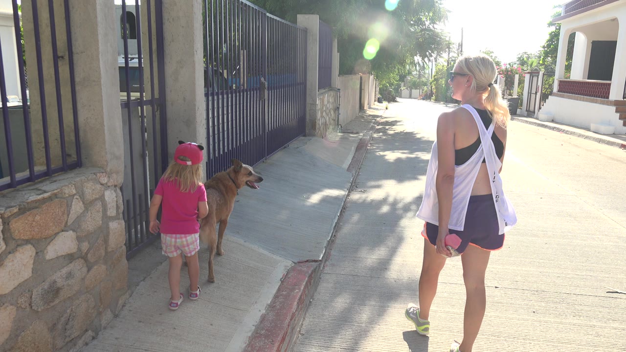 Woman, child, and tan dog walking beside a black metal gate on a sunny residential street.