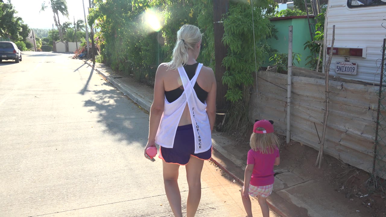 At 0:01, the woman and child are farther down the Grand Junction street, with a white trailer, a parked car, dense greenery, and bright lens flare visible in the background.