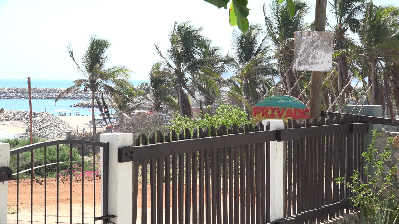 Opening frame of a brown metal gate and white post in Grand Junction, Colorado, with palm trees, a red private sign, pale sand, blue water, and no visible people.
