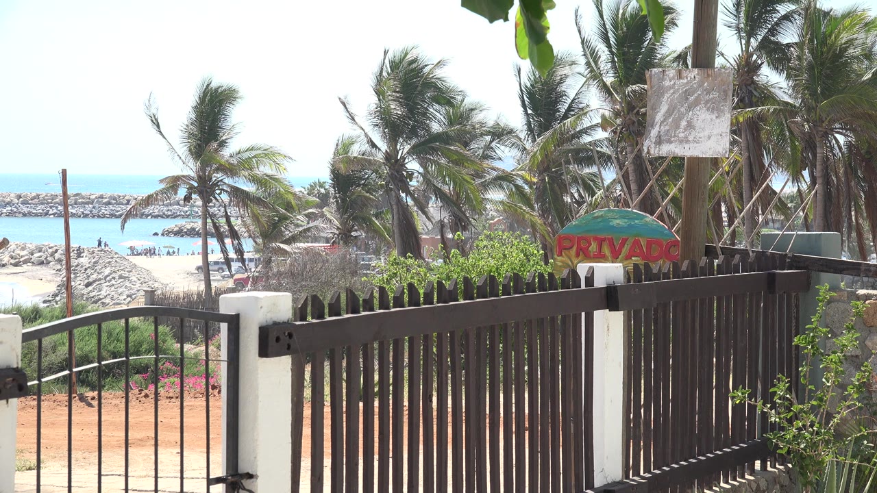 Opening frame of the Grand Junction 2017 clip showing a dark metal fence, white posts, palm trees, pale sand, rocky shoreline, blue water, and no clearly visible people.