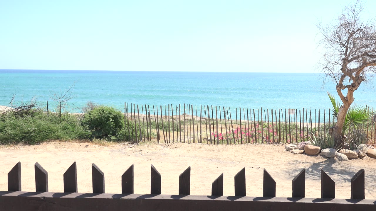At 0:01, the Grand Junction, Colorado frame holds on the sandy foreground, repeated dark fence posts, green shrubs, stick fencing, blue water horizon, and no visible people.