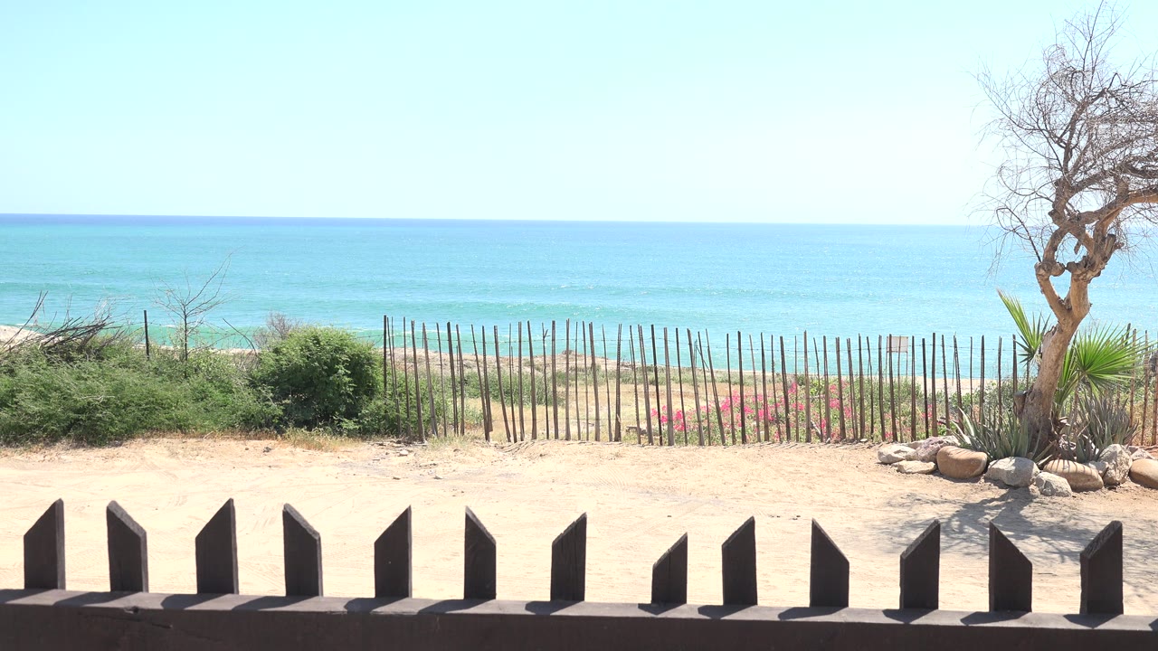 Opening frame of the Grand Junction Colorado clip showing pale sand, dark foreground posts, shrubs, a stick fence, turquoise ocean, bright sky, and no visible people.
