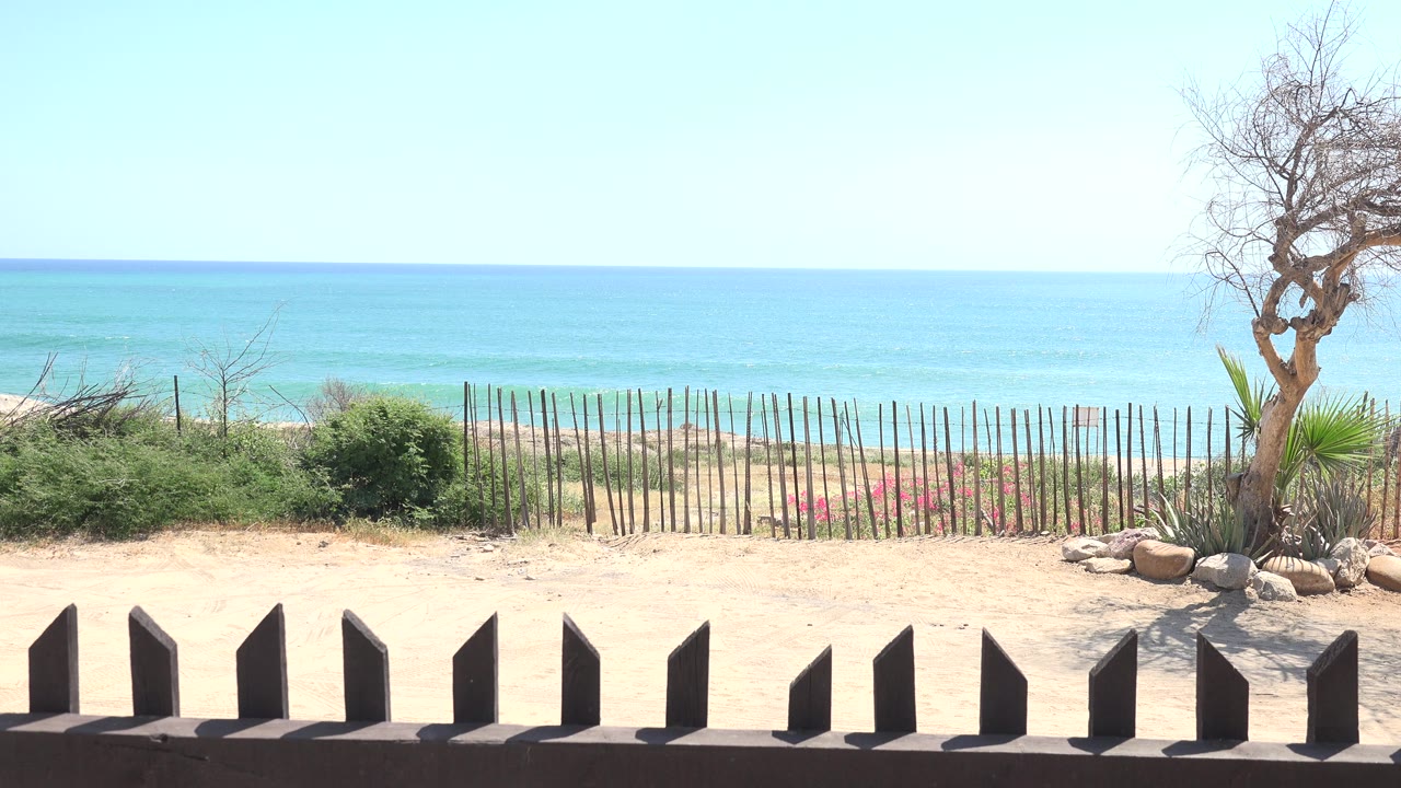 Second frame in Grand Junction Colorado shows the same empty beach overlook, with the bare tree near the right edge, low black fence posts below, and blue water across the horizon.