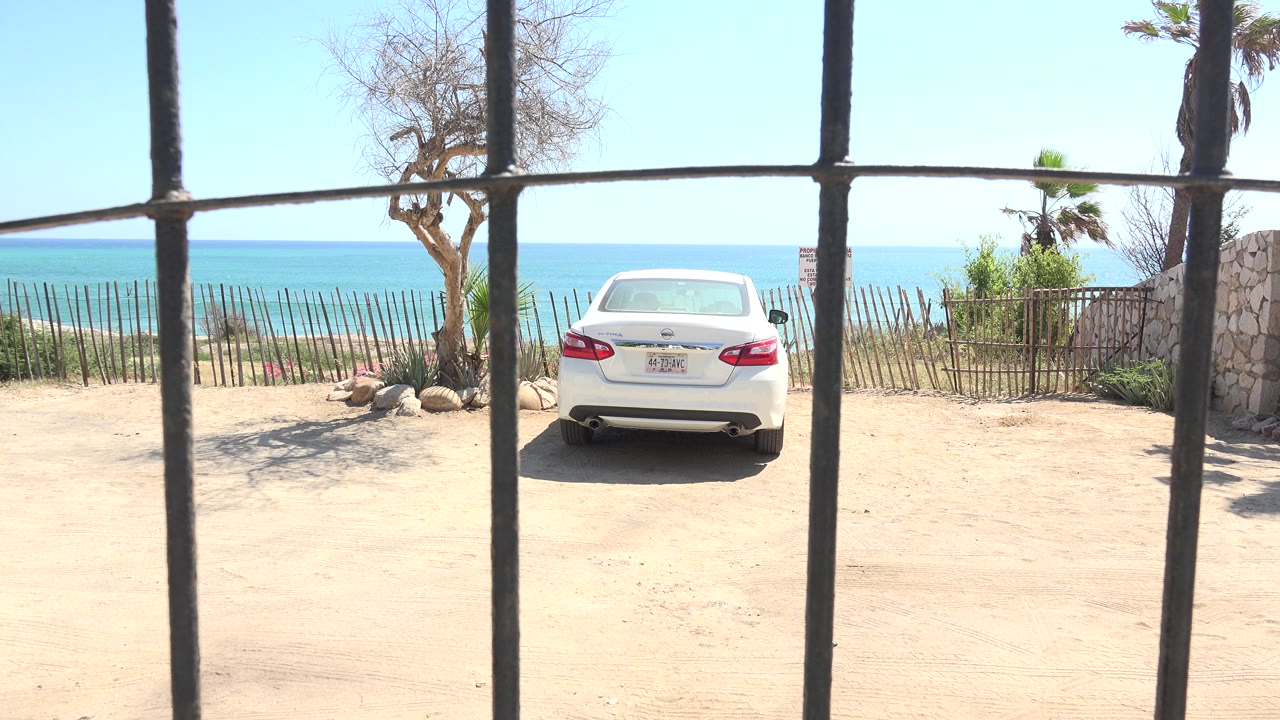 Parked white sedan framed by bars with sparse trees and blue horizon behind.