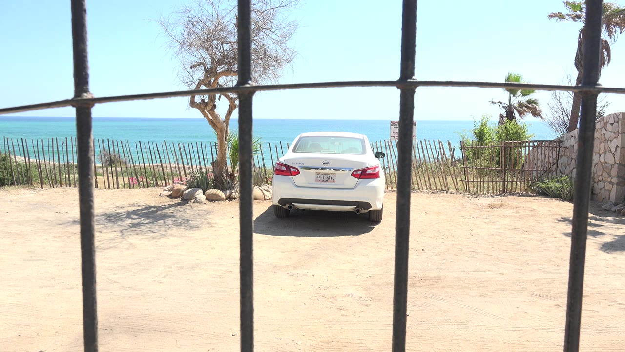 At 0:00, a white sedan is parked on a pale dirt surface in Grand Junction, Colorado, viewed through dark vertical bars with a low fence, trees, blue water, and no visible people.