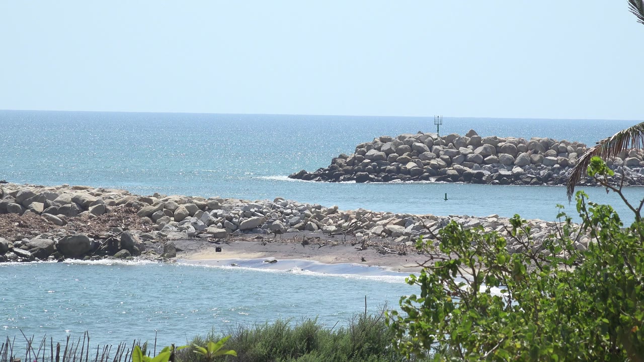 Blue ocean, rocky shore, breakwater, sky, and foreground greenery.