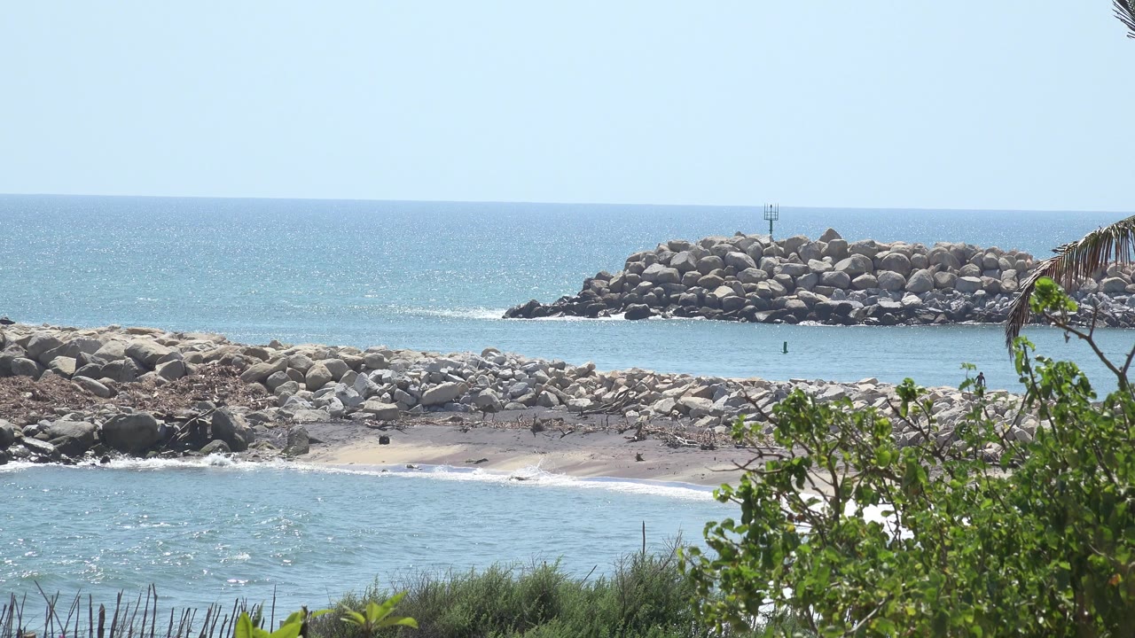 Opening frame of a Grand Junction, Colorado shoreline with blue water, a rocky sandbar, a distant rock breakwater, green foreground shrubs, and no visible people.