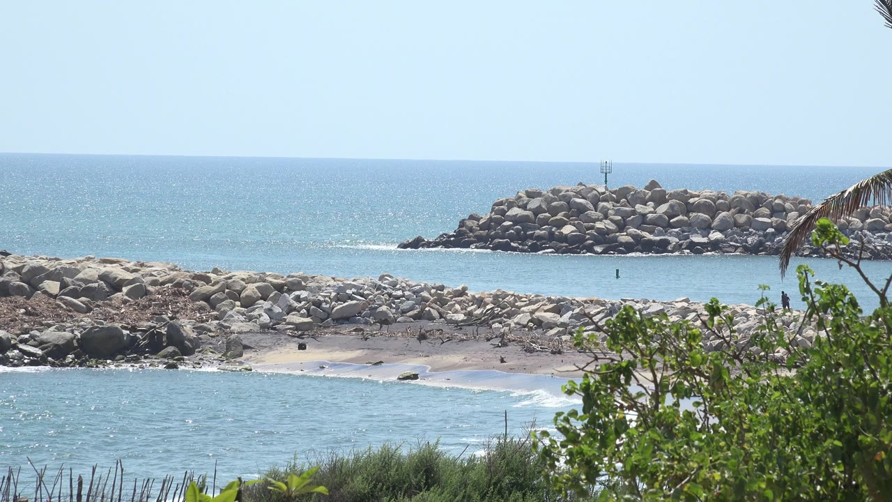 Opening frame of the Grand Junction, Colorado clip showing blue water in front of a rocky shore and distant rock breakwater, with green leaves at the right edge and no visible people.
