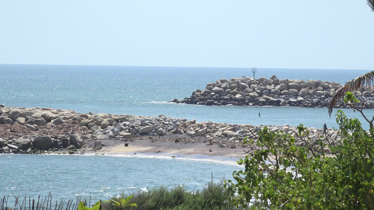 Rock breakwater behind a sandy rocky shoreline and blue water.