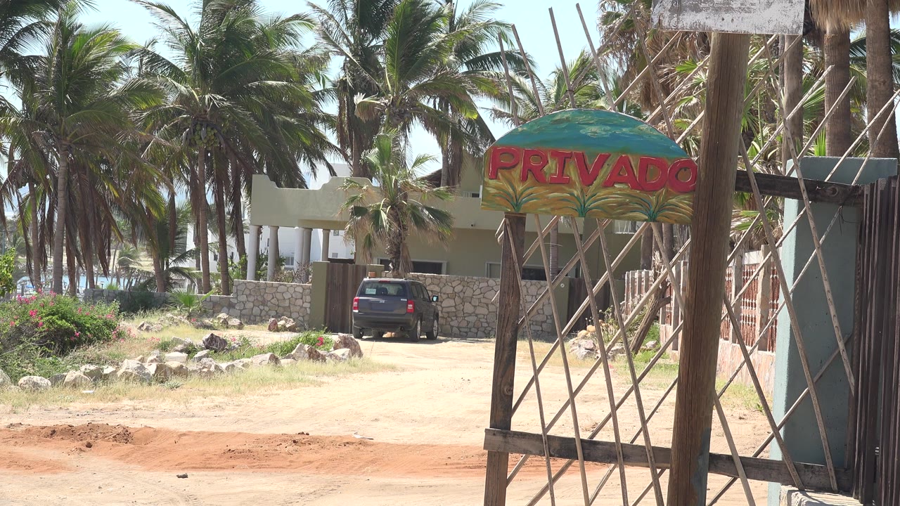 At 0:01, the metal gate and PRIVADO sign dominate the foreground while the parked car, low stone wall, pale building, and palms remain in bright daylight, with no visible people.