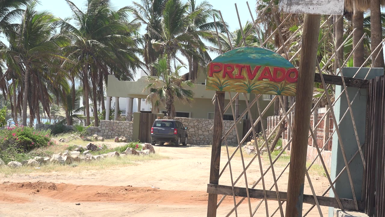 A Grand Junction, Colorado exterior frame shows a dirt road leading past a metal gate toward palm trees, a light building, a PRIVADO sign, and a dark parked SUV, with no visible people.