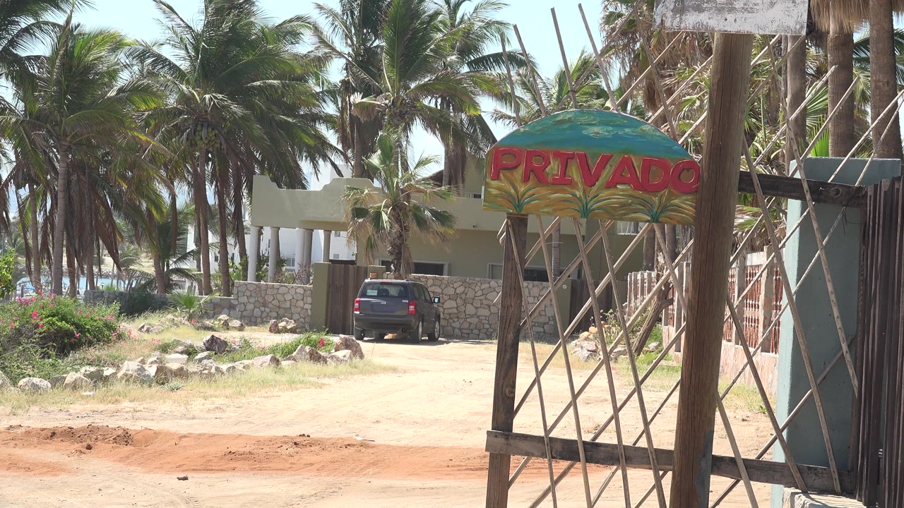 At 0:01 in Grand Junction, Colorado, the parked black car remains centered beyond the tan foreground, framed by gate posts, a stone wall, palm trees, and the PRIVADO sign, with no visible people.