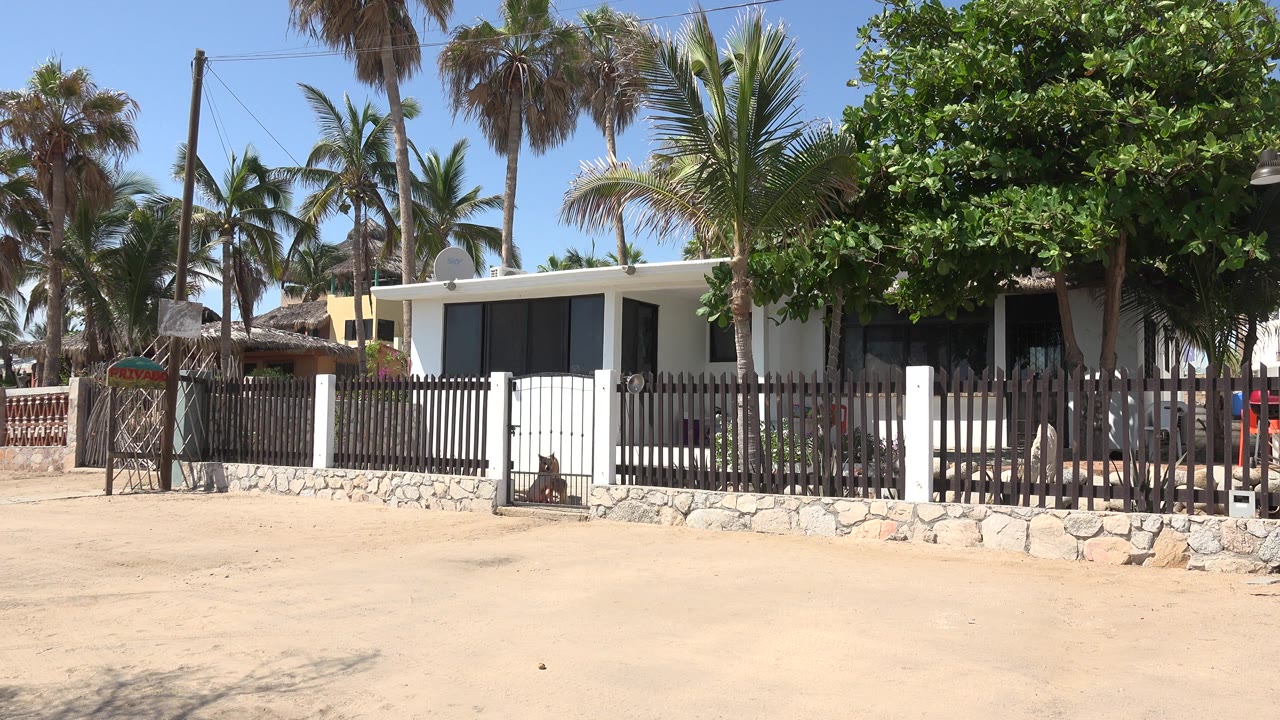 White house behind a brown fence with palms and sandy foreground.