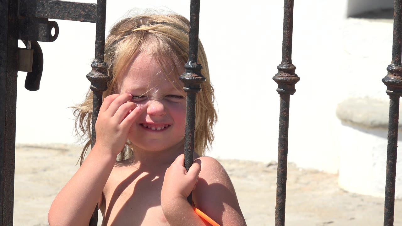 A young blonde girl in Grand Junction, Colorado squints in bright sun while touching her face behind dark iron gate bars.