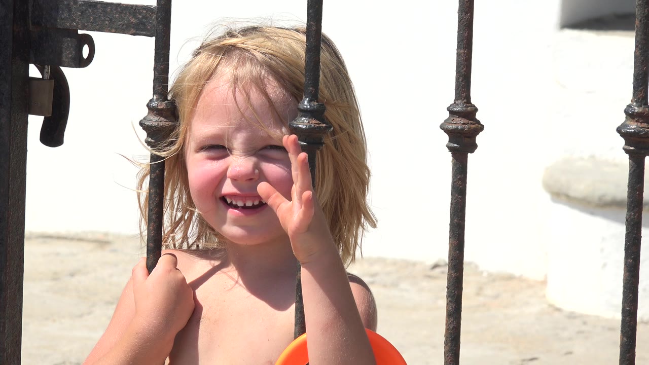 At 0:00 in Grand Junction, Colorado, a blond child smiles toward the camera while one hand rests on a black iron gate and the other lifts near her face.