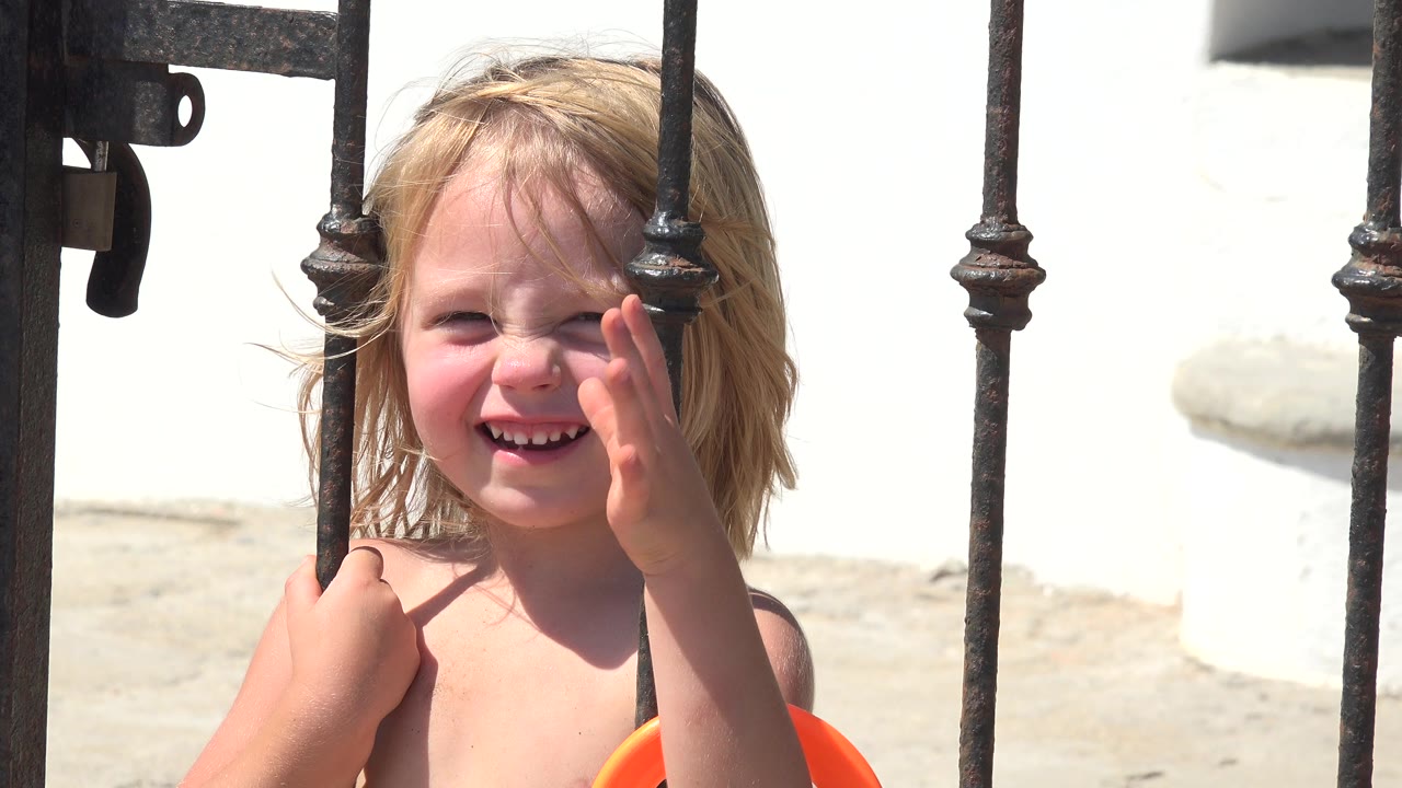 At 0:01 in Grand Junction, Colorado, the child keeps smiling behind the iron gate, with her raised hand closer to her cheek and bright light washing the white wall behind her.