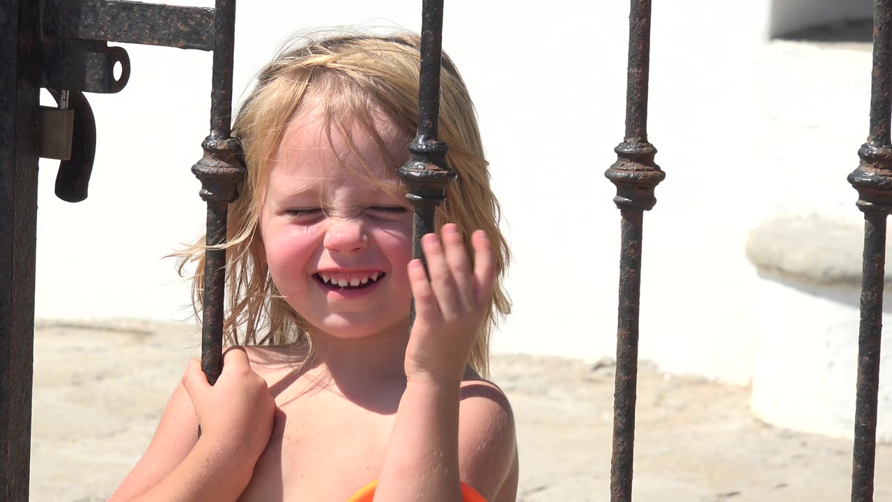 Child smiling as her hand moves across her face near an iron gate.