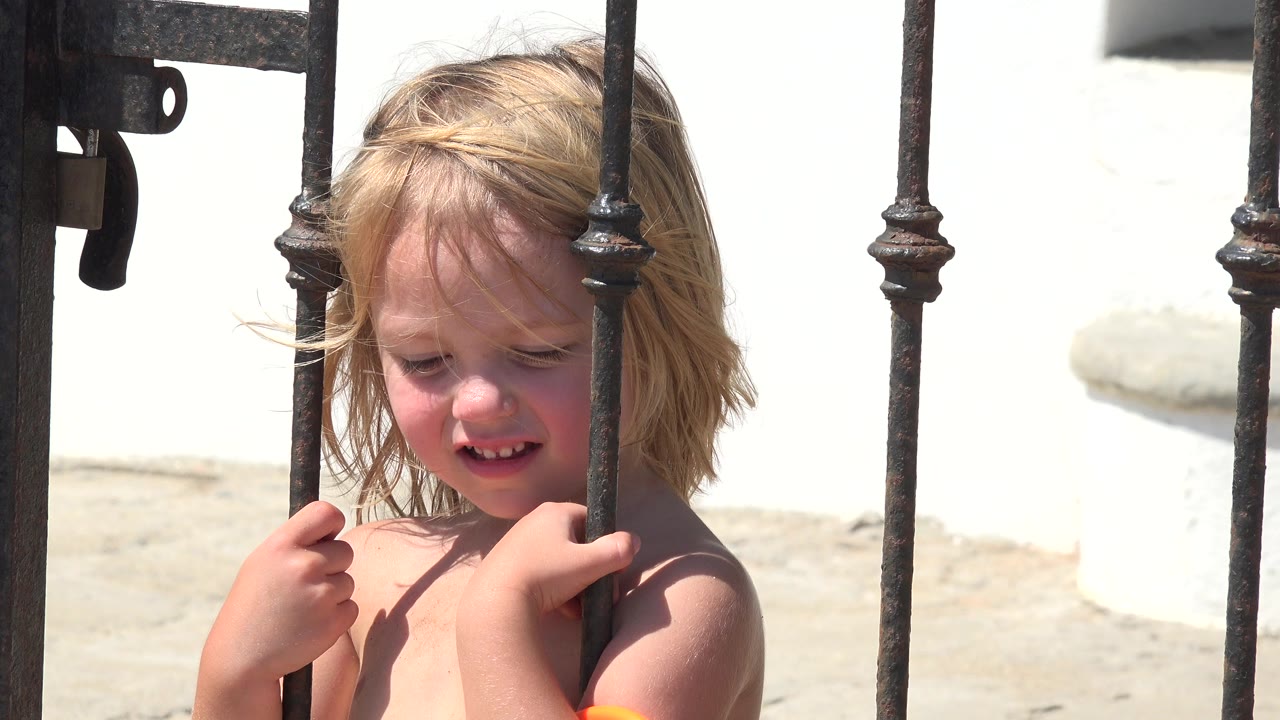 A blond child in Grand Junction, Colorado stands close behind black iron gate bars, looking downward with both hands near the metal in bright sun.